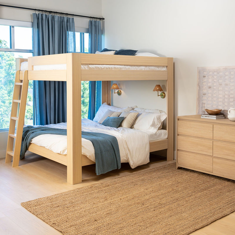 Modern light wood bunk bed with ladder in bright kids' bedroom, white bedding and blue throw, blue curtains, woven jute rug and wooden dresser