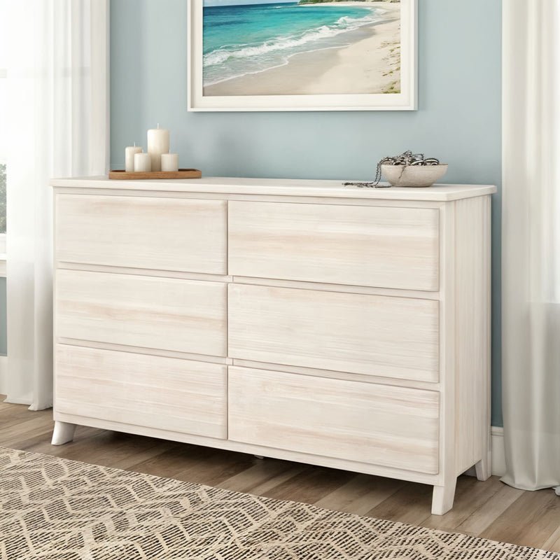 Whitewashed six-drawer wooden dresser in coastal-style bedroom with beach artwork, decorative candles, and light wood flooring