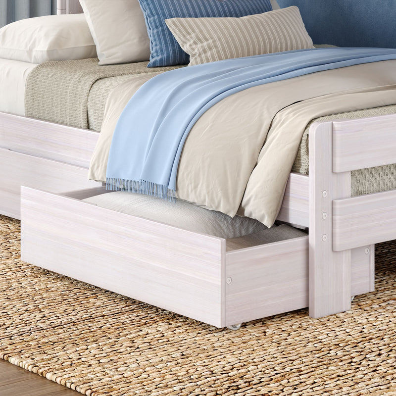Whitewashed wooden trundle bed with beige bedding, light blue throw and striped pillows on woven jute rug, showing under-bed storage drawer