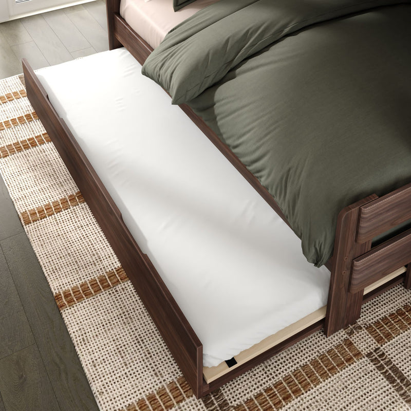 Walnut wooden trundle bed drawer pulled out with white mattress beneath a bed with green duvet on a patterned rug — space-saving underbed storage for small bedrooms