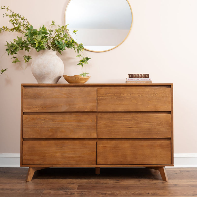 Mid-century modern six-drawer wooden dresser in warm oak on hardwood floor with round wall mirror, ceramic vase of green foliage, wooden bowl and stacked books against a neutral beige wall
