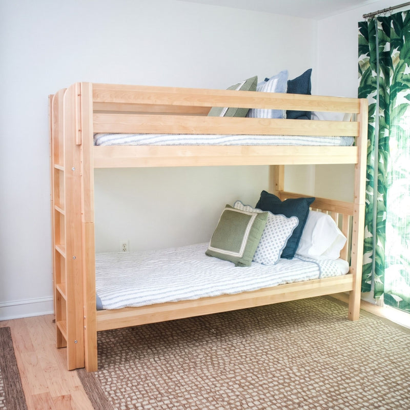 A light natural wood bunk bed with a ladder on the left, styled with striped bedding and decorative pillows in a bright bedroom with a patterned rug and green leaf-print curtains.