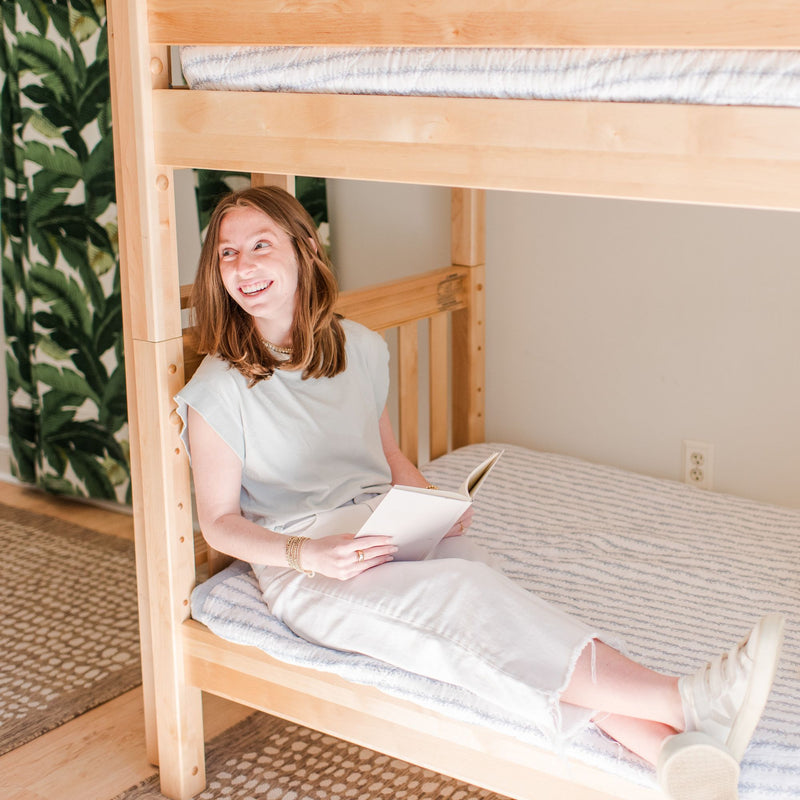 Smiling woman reading a book on a light-wood bunk bed in a bright bedroom with tropical leaf wallpaper