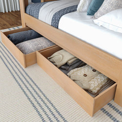 Underbed storage drawers in light wood platform bed filled with folded sweaters, blankets and decorative pillows on striped rug — space-saving bedroom storage solution