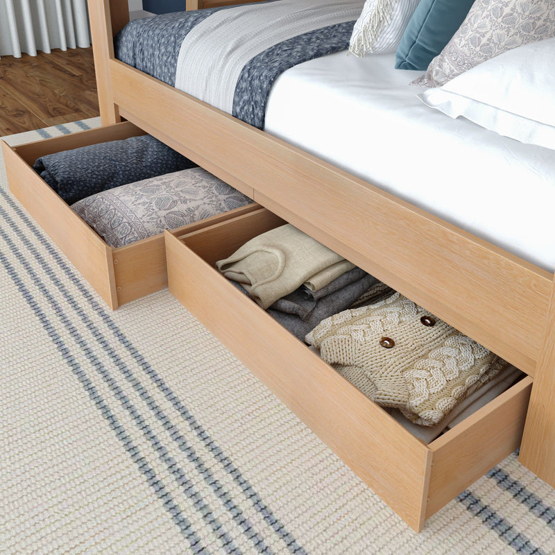 Underbed storage drawers in light wood platform bed filled with folded sweaters, blankets and decorative pillows on striped rug — space-saving bedroom storage solution