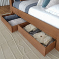 Wooden platform bed with open under-bed storage drawers holding folded sweaters, blankets and pillows — space-saving bedroom storage solution.