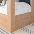 Close-up of a light oak four-poster bed frame corner with crisp white bedding, decorative pillows and a striped woven rug in a modern farmhouse bedroom