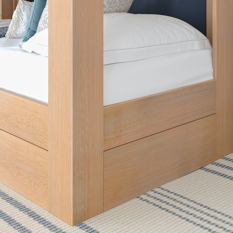Close-up of a light oak four-poster bed frame corner with crisp white bedding, decorative pillows and a striped woven rug in a modern farmhouse bedroom