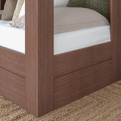 Close-up of walnut-finish four-poster platform bed corner with beige bedding, low-profile mattress and integrated storage panel on woven jute rug in a modern farmhouse bedroom