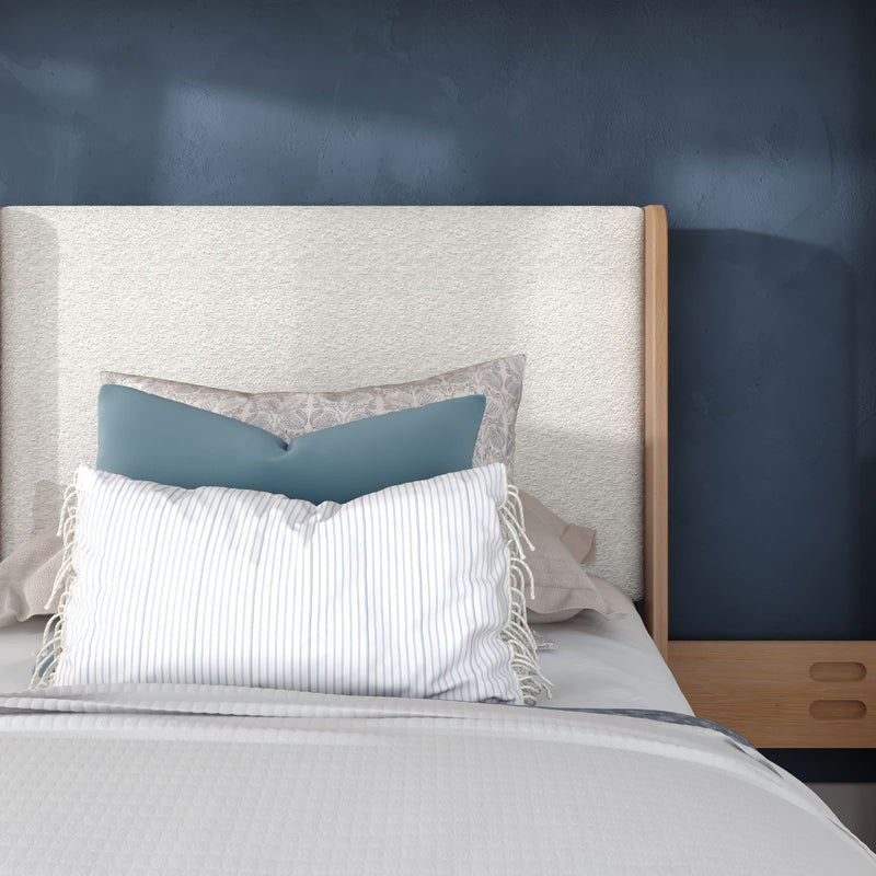 Modern bedroom close-up of a bed with a neutral upholstered headboard, layered blue and striped decorative pillows, white textured bedding and a wooden nightstand against a navy accent wall.