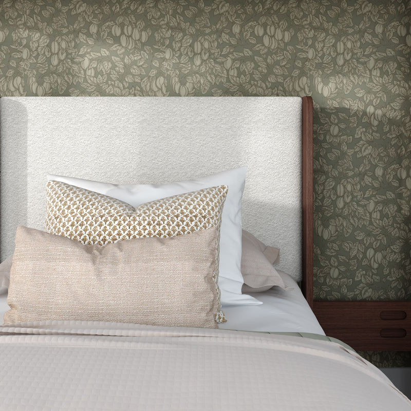 Cozy modern bedroom close-up showing a white upholstered headboard, layered neutral and patterned throw pillows, textured beige bedding, green botanical wallpaper, and a wooden bedside table