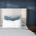 Cozy modern bedroom with upholstered light beige headboard, layered decorative pillows (blue lumbar, patterned and striped), navy accent wall and wooden nightstand