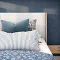 Navy blue bedroom with light gray upholstered headboard, wooden bed frame, layered decorative pillows (blue velvet, black-and-white floral, striped fringed lumbar) and blue floral quilt