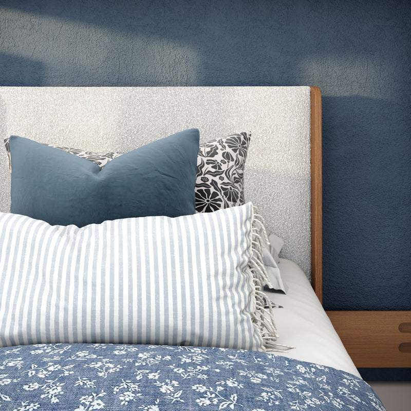 Close-up of a modern coastal bedroom with a textured upholstered headboard, blue floral quilt, striped lumbar pillow and decorative navy and patterned throw pillows on a wooden bed frame