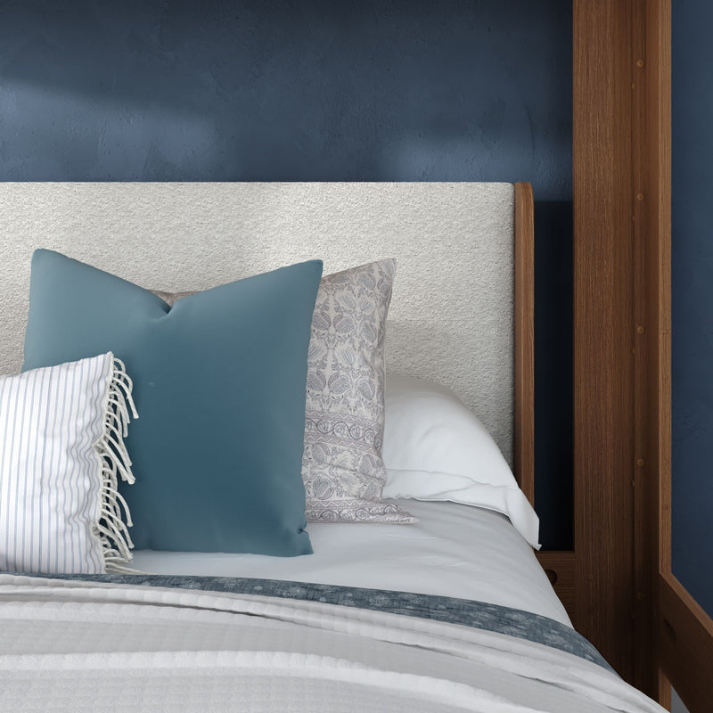 Close-up of a modern bedroom bed with a textured light-gray upholstered headboard, navy blue accent wall and wooden frame, white linen bedding and layered decorative pillows including a teal square pillow, patterned neutral pillow and striped fringed pillow