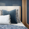 Close-up of cozy bedroom decor with navy blue decorative pillows, striped and floral accent pillows, textured cream upholstered headboard, wooden bed frame and blue floral quilt