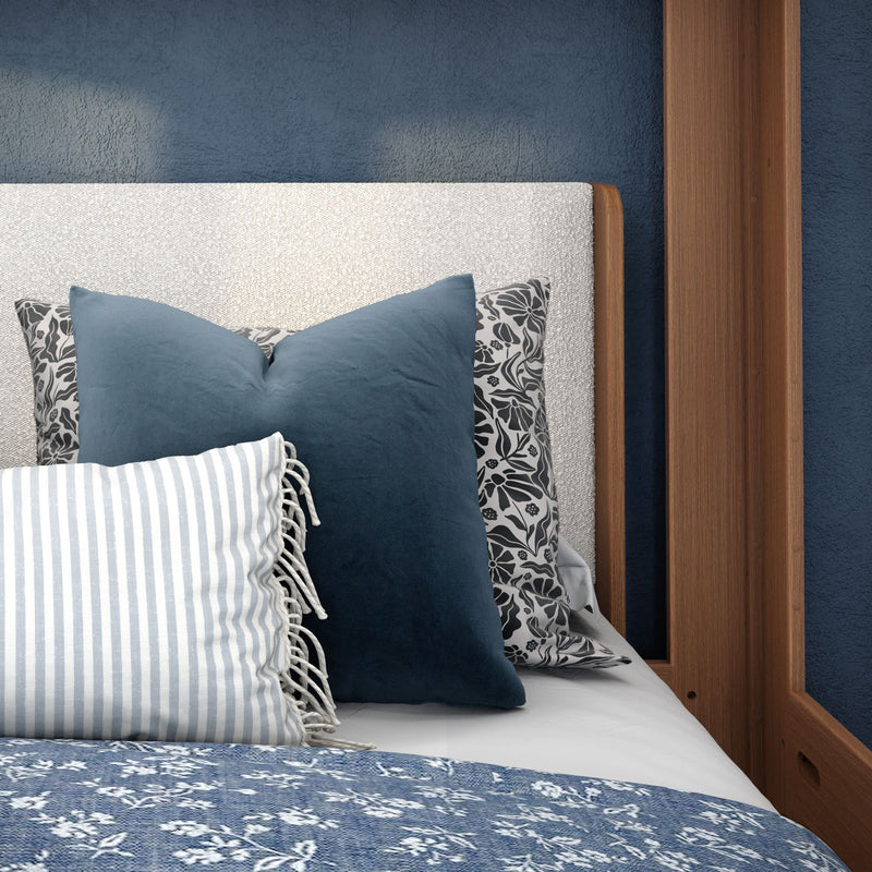 Blue and white bedroom decor with navy velvet throw pillows, striped fringed lumbar pillow, floral pillowcases, textured upholstered headboard and wooden bed frame against a navy blue wall
