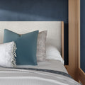 Close-up of a modern bedroom with a white textured headboard, navy accent wall, wooden bed frame and blue patterned throw pillows on white bedding