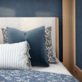 Close-up of cozy coastal bedroom decor with navy velvet throw pillow, striped fringed lumbar pillow, floral accent pillow, textured cream headboard, blue floral quilt and light oak bed frame