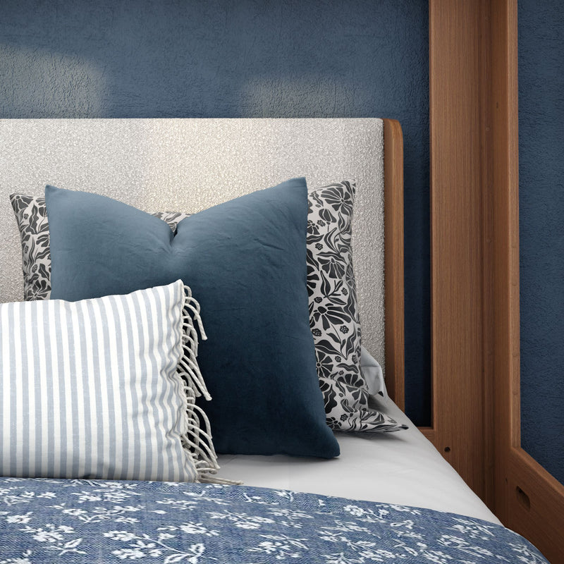 Cozy bedroom close-up of navy velvet throw pillows, striped fringe lumbar pillow and black-and-white floral cushion on a textured light-gray upholstered headboard with wooden bed frame and navy accent wall
