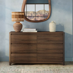 Walnut six-drawer wooden dresser in modern bedroom styled with woven table lamp, ceramic vase, stacked books and round mirror reflecting red rock canyon