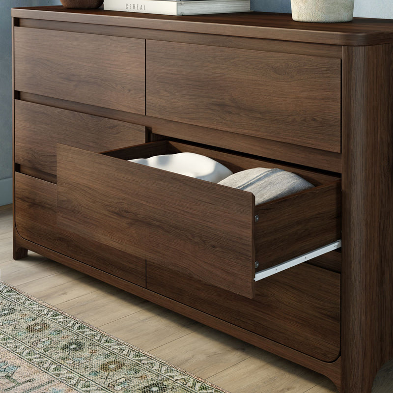 Walnut-finish wooden dresser with an open bottom drawer showing folded bedding and pillow, modern bedroom storage chest on hardwood floor with patterned rug