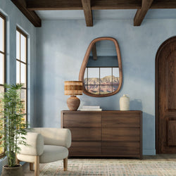 Modern boho living room with blue plaster walls and rustic wood beams, walnut dresser with oval wooden mirror reflecting a desert landscape, wicker table lamp and ceramic vase, neutral beige lounge chair and potted plant by large windows with natural light