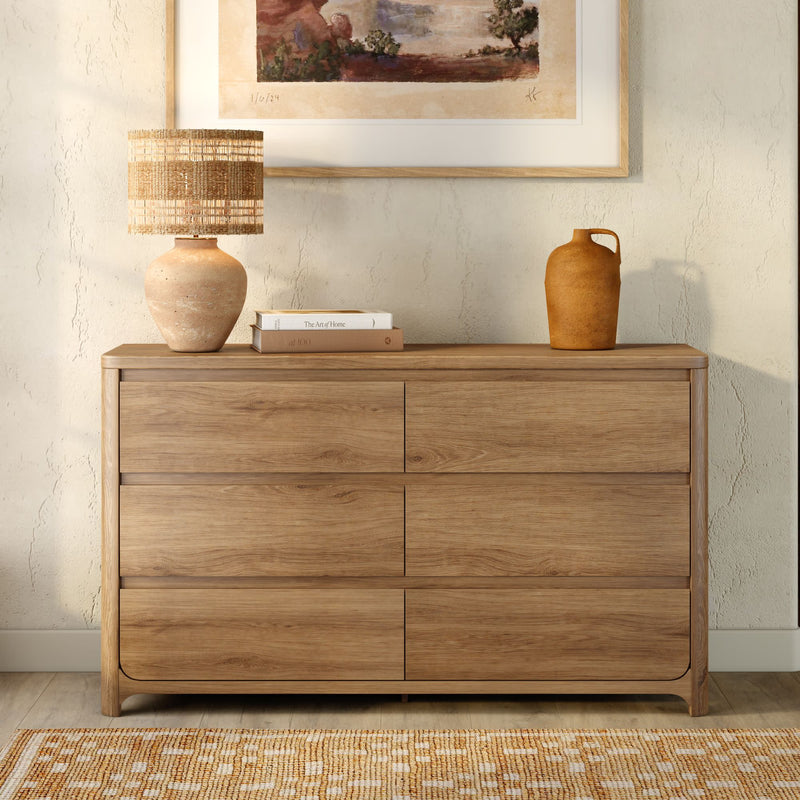 Modern six-drawer oak dresser with woven table lamp, stacked books and ceramic vase beneath framed wall art in neutral home decor