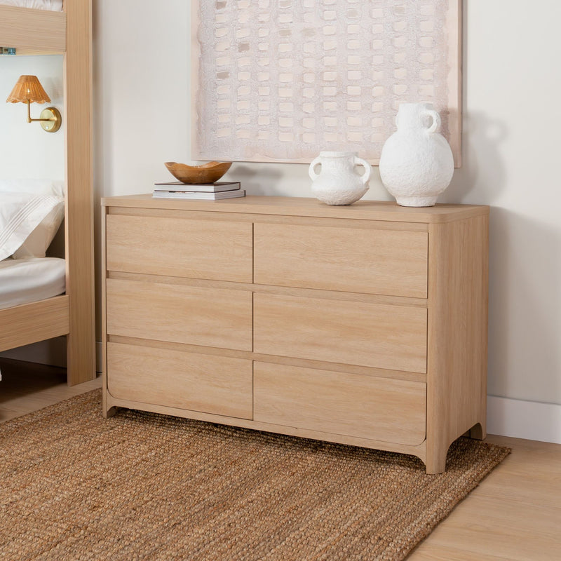 Light oak dresser in minimalist Scandinavian bedroom decor with textured white ceramic vases, wooden bowl on stacked books, beige abstract wall art, brass sconce and natural jute rug