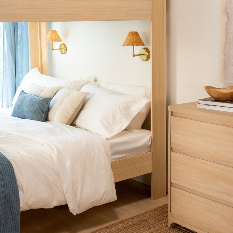 Cozy Scandinavian bedroom with white bedding, layered neutral pillows, light wood canopy bed frame, rattan wall sconces and blue curtains