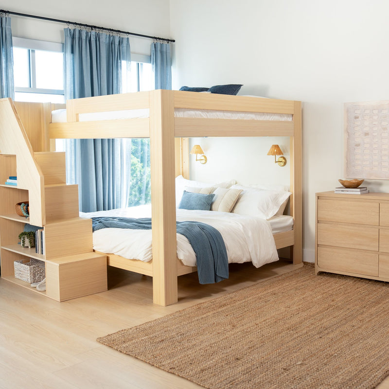 Natural wood bunk bed with built-in staircase storage, white bedding and blue throw, blue curtains and brass wall sconces in a bright kids' bedroom