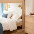 Cozy Scandinavian bedroom with light wood bed frame, crisp white bedding, blue accent pillows, rattan wall sconces and natural wood dresser