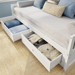 White twin daybed with two open underbed storage drawers containing folded blankets and pillows on a woven rug — modern space-saving bedroom storage solution