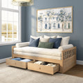 Light wood daybed with under-bed storage drawers, cream mattress and blue and green throw pillows in a cozy blue-gray living room with framed watercolor wall art and a large window letting in natural light