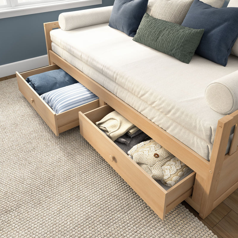Modern wooden daybed with built-in underbed storage drawers open, cream mattress and bolsters, blue and green decorative pillows on a textured beige rug, space-saving bedroom furniture