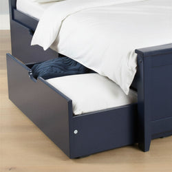 Navy blue trundle bed with pull-out storage drawer containing a white mattress and navy knitted blanket on light hardwood floor