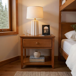 Wooden nightstand with a modern lamp, glass, and framed photo beside a cozy bed in a stylish bedroom setting.