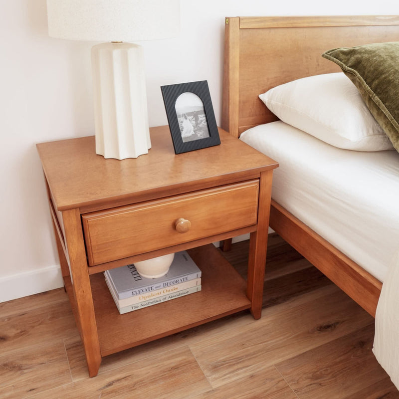 Wooden nightstand with a drawer, featuring a white table lamp, a photo frame, and stacked books, next to a bed with green and white bedding.