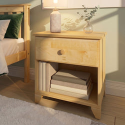 Light wood nightstand with patterned ceramic bedside lamp, glass vase of eucalyptus, stacked books and green pillow in a cozy minimalist bedroom bathed in warm sunlight