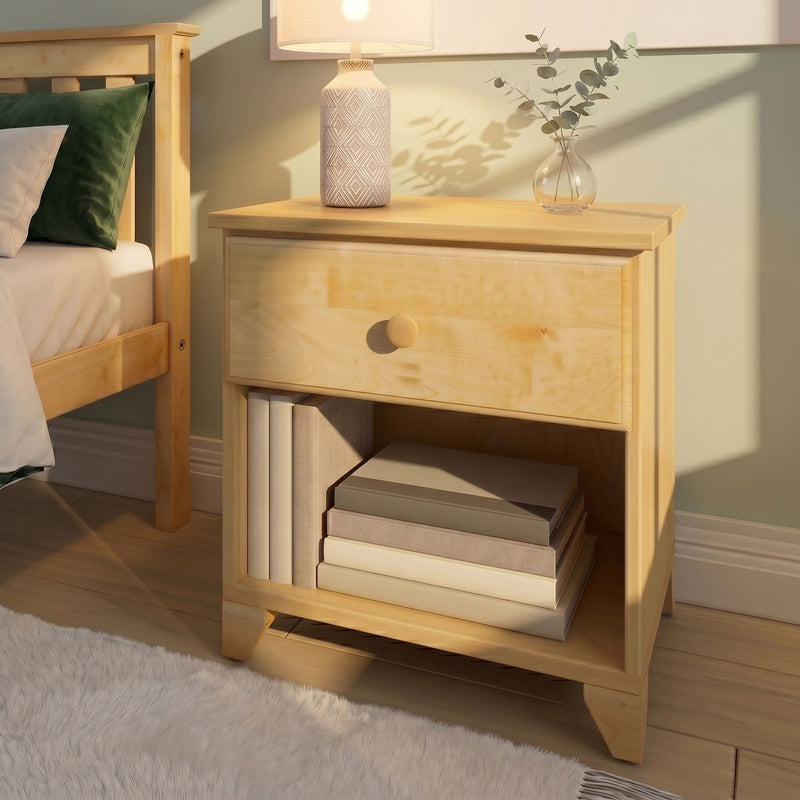 Light wood nightstand with patterned ceramic bedside lamp, glass vase of eucalyptus, stacked books and green pillow in a cozy minimalist bedroom bathed in warm sunlight