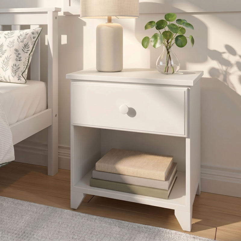 White nightstand with ceramic table lamp, glass vase holding Pilea peperomioides (Chinese money plant), stacked books and botanical pillow in neutral bedroom decor