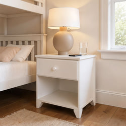 White nightstand with lamp and glass near bed in cozy bedroom setting.