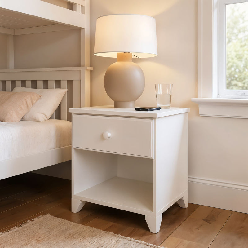 White nightstand with lamp and glass near bed in cozy bedroom setting.