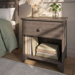 Wooden bedside table with beige ceramic lamp, glass vase of green leaves and stacked books in a cozy bedroom