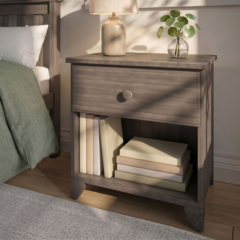 Wooden bedside table with beige ceramic lamp, glass vase of green leaves and stacked books in a cozy bedroom