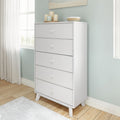 Modern white tall five-drawer dresser with angled mid-century legs in a bright bedroom with pastel blue wall, hardwood floor, window light and decorative candles