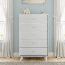 Modern white 5-drawer wooden dresser against a light blue wall with candles and a bowl on top, flanked by white curtains in a coastal-style bedroom