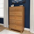Mid-century modern tall five-drawer oak dresser chest of drawers in natural wood finish against a navy blue accent wall, styled in a bedroom with white curtains and hardwood floor