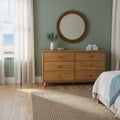 Modern coastal bedroom with natural wood six-drawer dresser, round rattan mirror, green accent wall and ocean view through sheer curtains