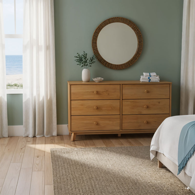 Modern coastal bedroom with natural wood six-drawer dresser, round rattan mirror, green accent wall and ocean view through sheer curtains